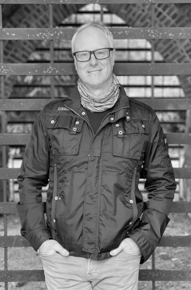 Man wearing a dark jacket and glasses standing in front of a wooden fence.